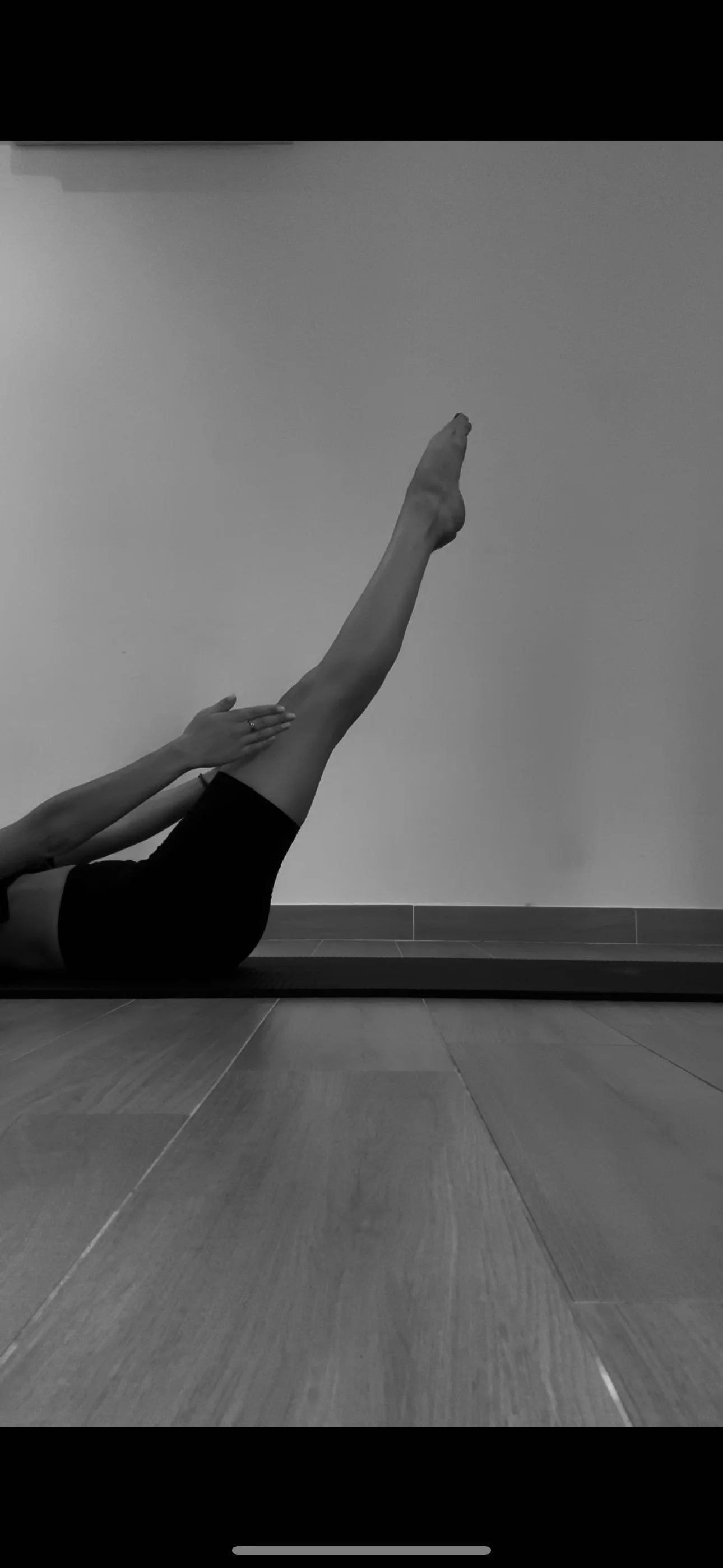 Une personne effectuant un étirement de jambes lors d'une séance de yoga ou de Pilates, allongée sur un tapis en bois, en salle ou studio.
