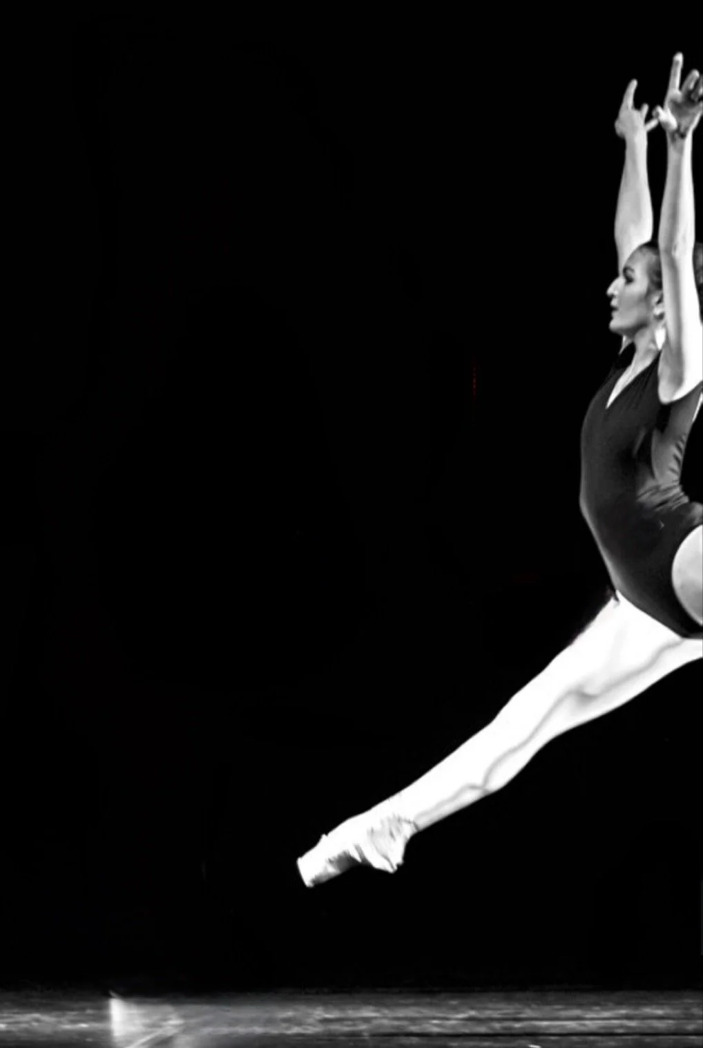 Une danseuse classique en plein saut, portant un justaucorps noir, dans un fond noir, en position de grand jeté avec les bras levés au-dessus de la tête.