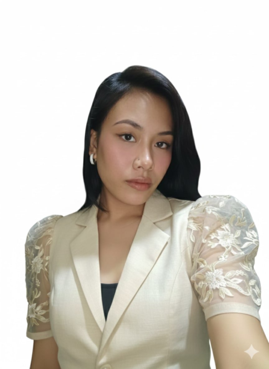 A woman with dark hair and red lipstick standing outdoors near a curved metal fence with green trees in the background.