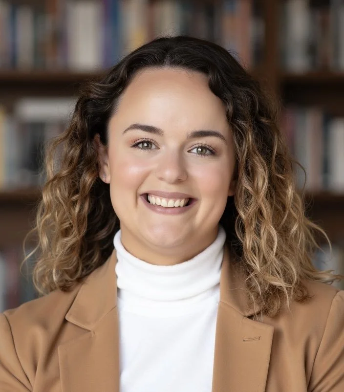 A young woman with curly brown hair smiling, wearing a beige blazer and white turtleneck, in front of a bookshelf.