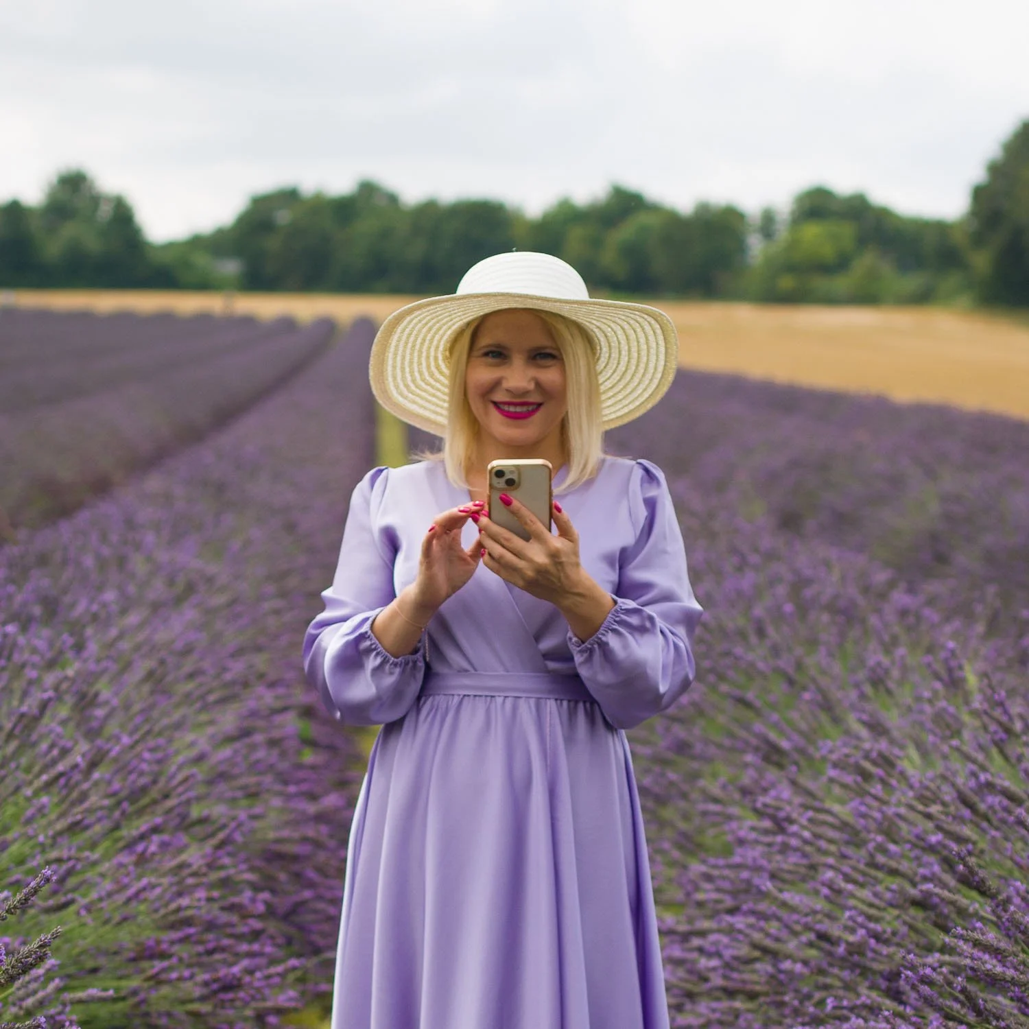 Oksana Pal steht im Fliederfeld, hält ihr Handy in beiden Händen und blickt strahlend in die Kamera – eine einladende Geste, um den ersten Schritt zu machen und gemeinsam Klarheit zu schaffen sowie Lösungen für deine Herausforderungen zu finden.