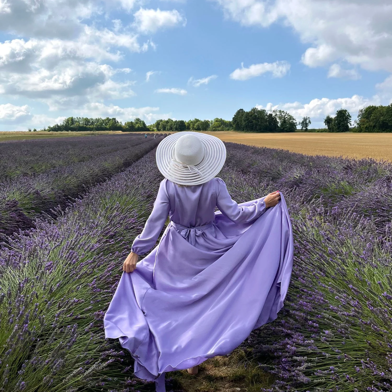 Eine Frau in einem lila Kleid und Sonnenhut rennt fröhlich durch ein Fliederfeld – ein Bild, das die Leichtigkeit und Freiheit symbolisiert, die durch eine individuelle Begleitung per Videoanruf oder Telefonat erreicht werden kann.