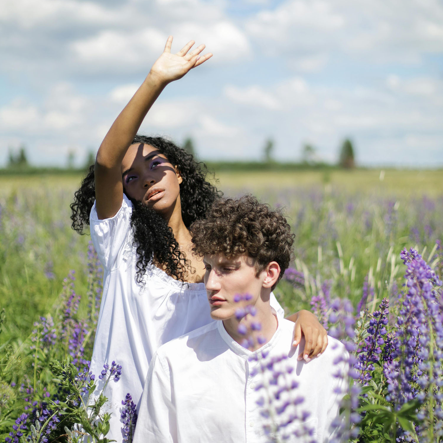 Eine Frau und ein Mann sitzen in einem Fliederfeld. Die Frau streckt ihre Hand zum Sonnenschein, während sie nach oben blickt – ein Symbol für das Streben nach innerer Klarheit und der Entfaltung des eigenen Potenzials.
