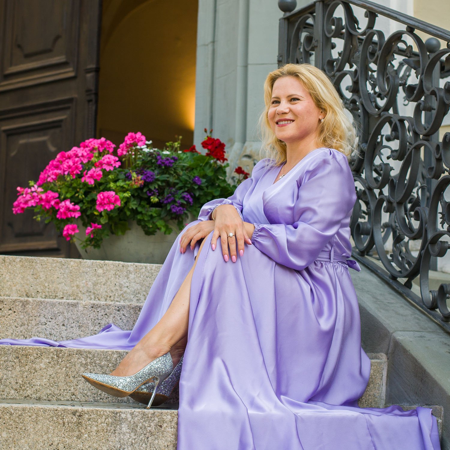 Oksana Pal von Freiwiederwind sitzt lächelnd in einem lila Kleid auf einer Treppe, umgeben von bunten Blumen – ein Symbol für die unterstützende und positive Philosophie ihrer Arbeit.