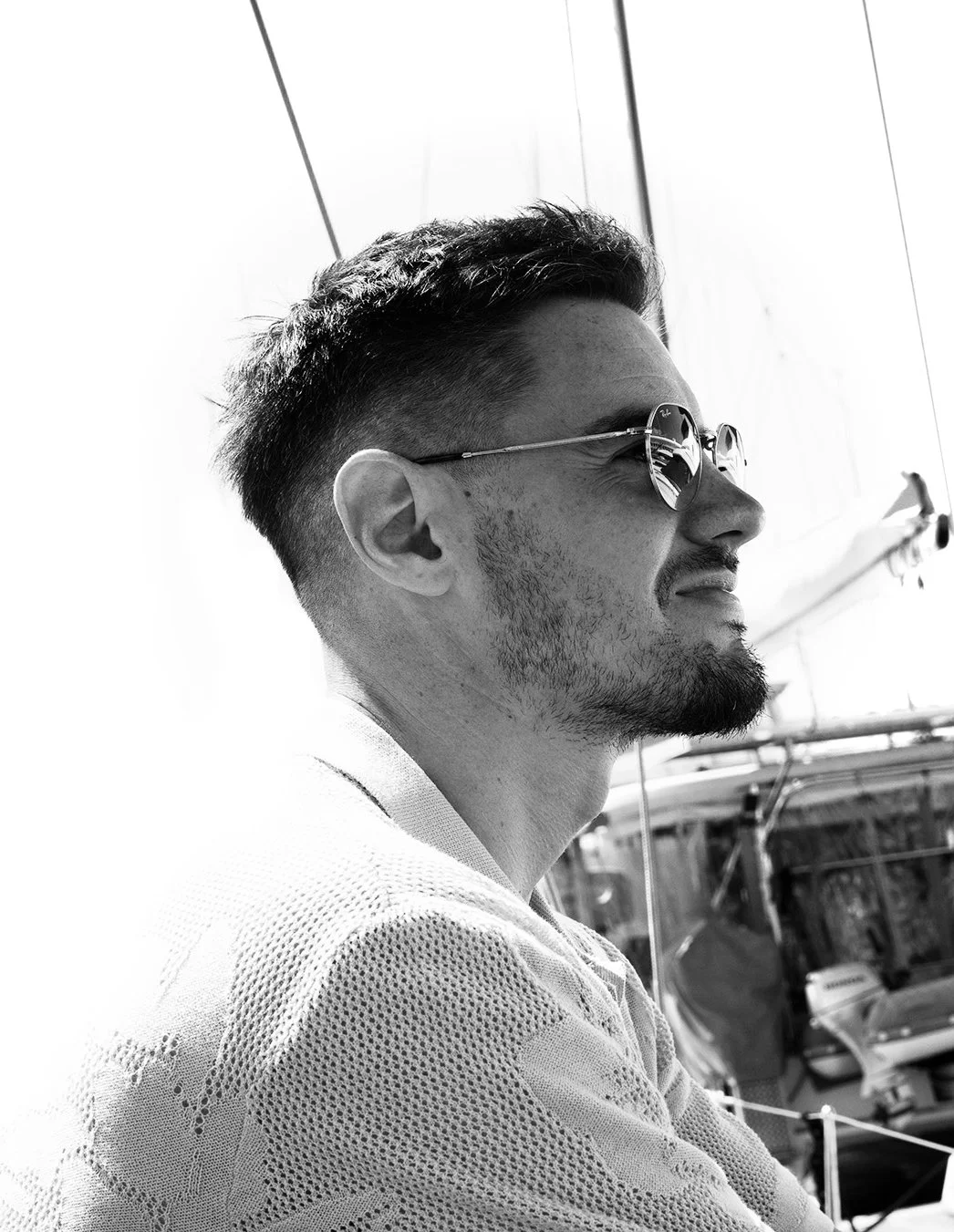 A black-and-white photo of a man in sunglasses and a textured shirt, looking to the side, with boats and strings of a sailboat in the background.