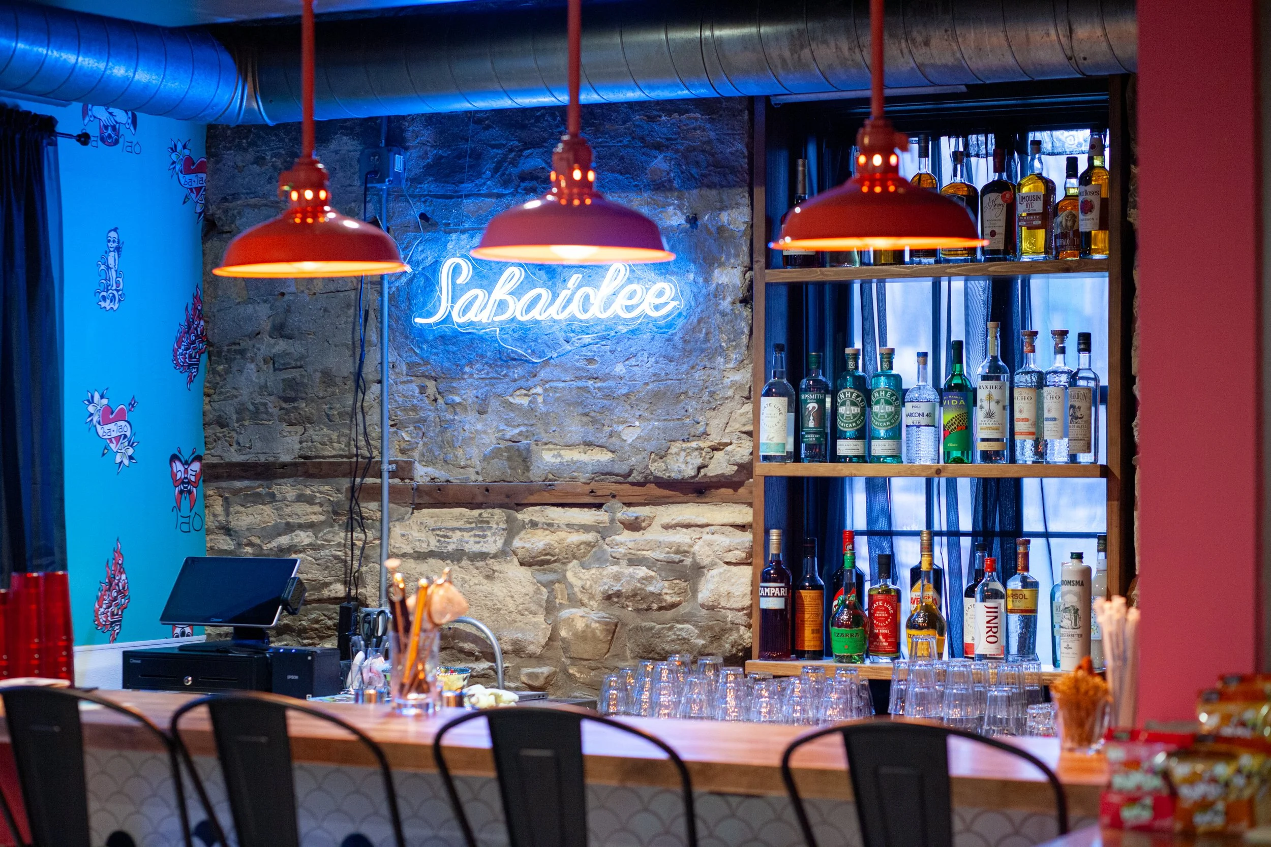 Interior view of Ba Lao Bar in Madison, Wisconsin featuring a blue neon “Sabaidee” sign mounted on an exposed stone wall, red pendant lights above a wood bar top, and open shelving displaying liquor bottles.