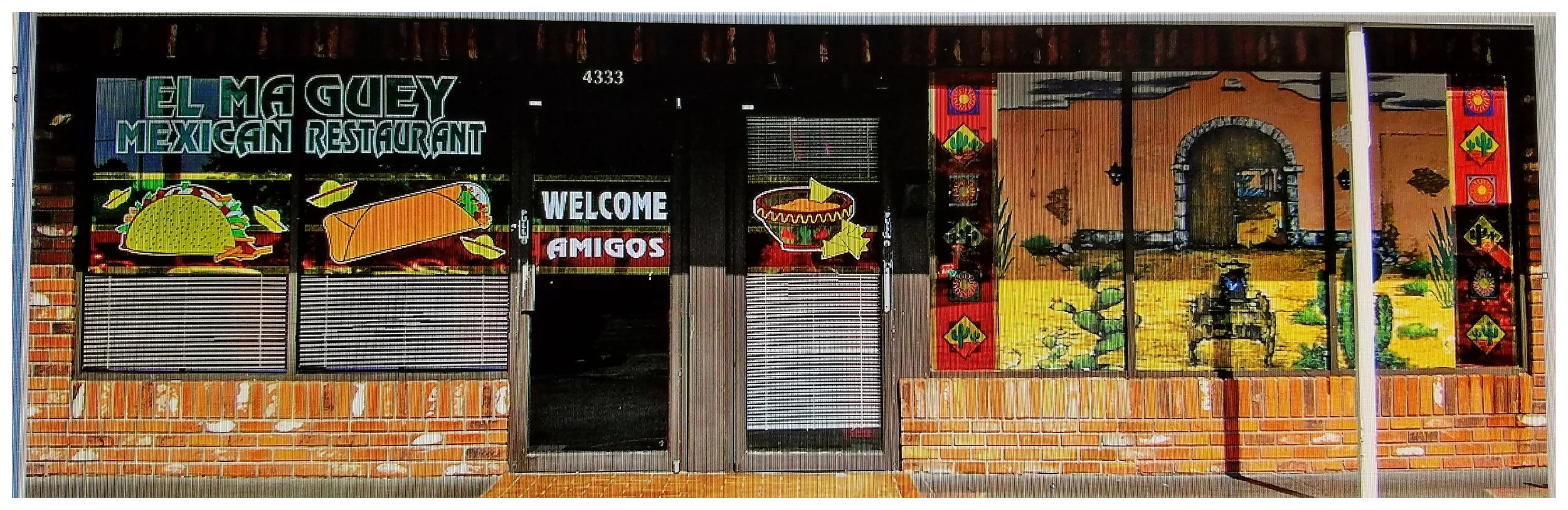 Exterior of El Maguey Mexican Restaurant with colorful window decorations featuring a taco, burrito, sombrero, and a painted scene of a Mexican village. Sign reads 'Welcome Amigos.' The building has brick and glass facade.