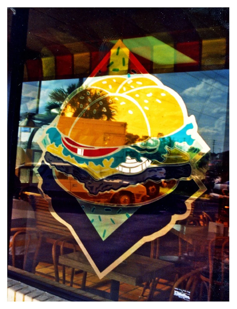 Colorful stained glass window depicting a burger with yellow buns, red tomato, green lettuce, and various layers, with reflections of palm trees and outdoor scenery.