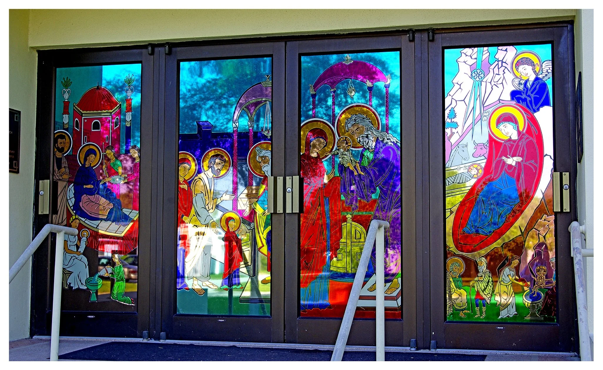 Stained glass doors depicting biblical scenes with halo-adorned figures, likely religious icons, on a building entrance.