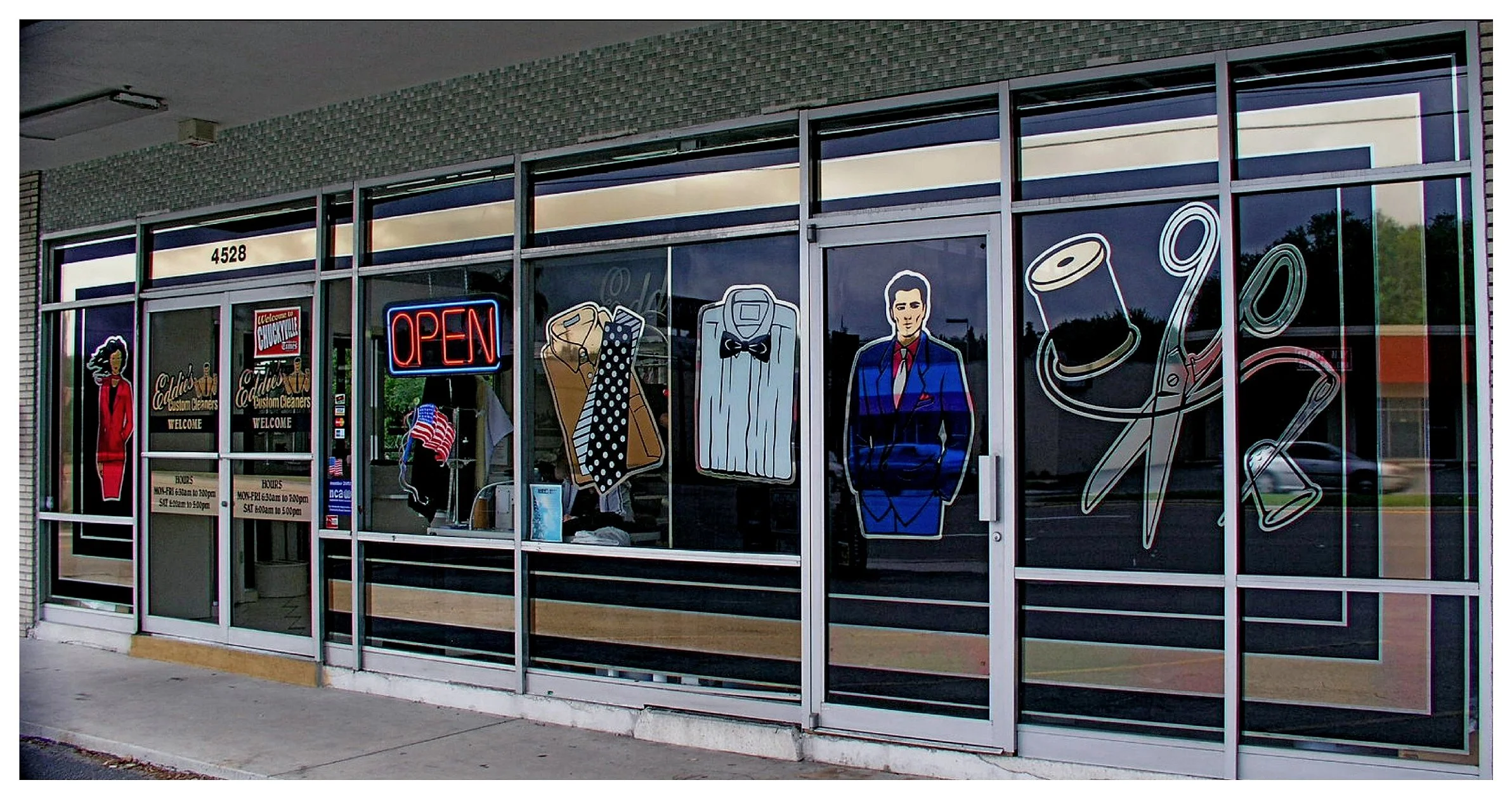 Dry cleaning storefront with large window graphics, including illustrations of clothing, scissors, and sewing thread, and a neon 'open' sign. colored window film