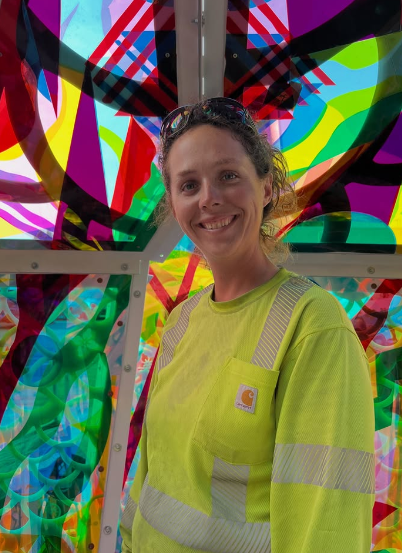 A smiling woman wearing a yellow safety shirt standing in front of colorful, abstract, graffiti-like artwork on a wall. colored window film
