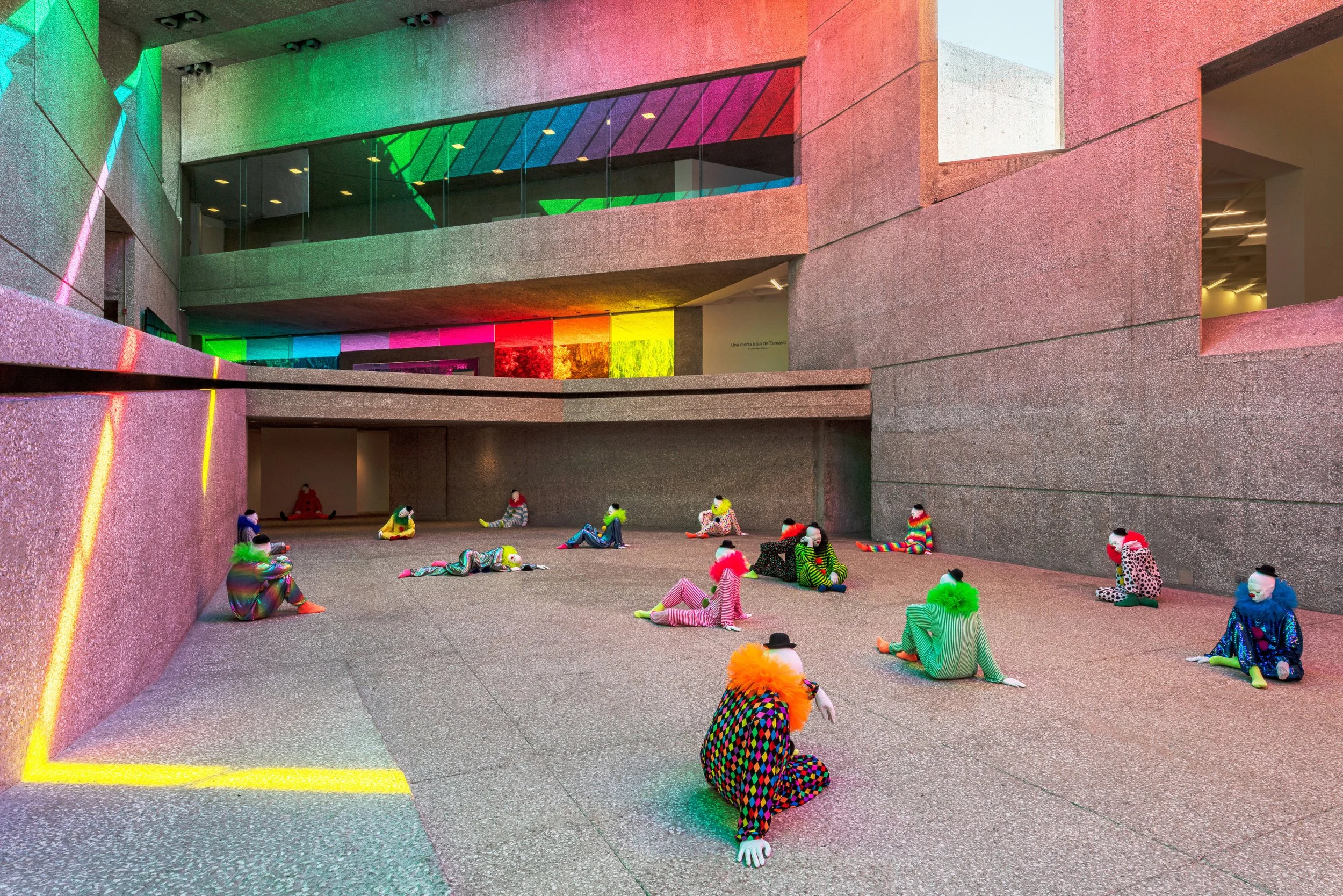 An art installation in a modern architectural space with concrete walls and floors, featuring numerous mannequins dressed in colorful, patterned clothing and wigs, sitting or lying on the ground. The background includes a large window with multicolored light reflections and a gallery or museum setting.