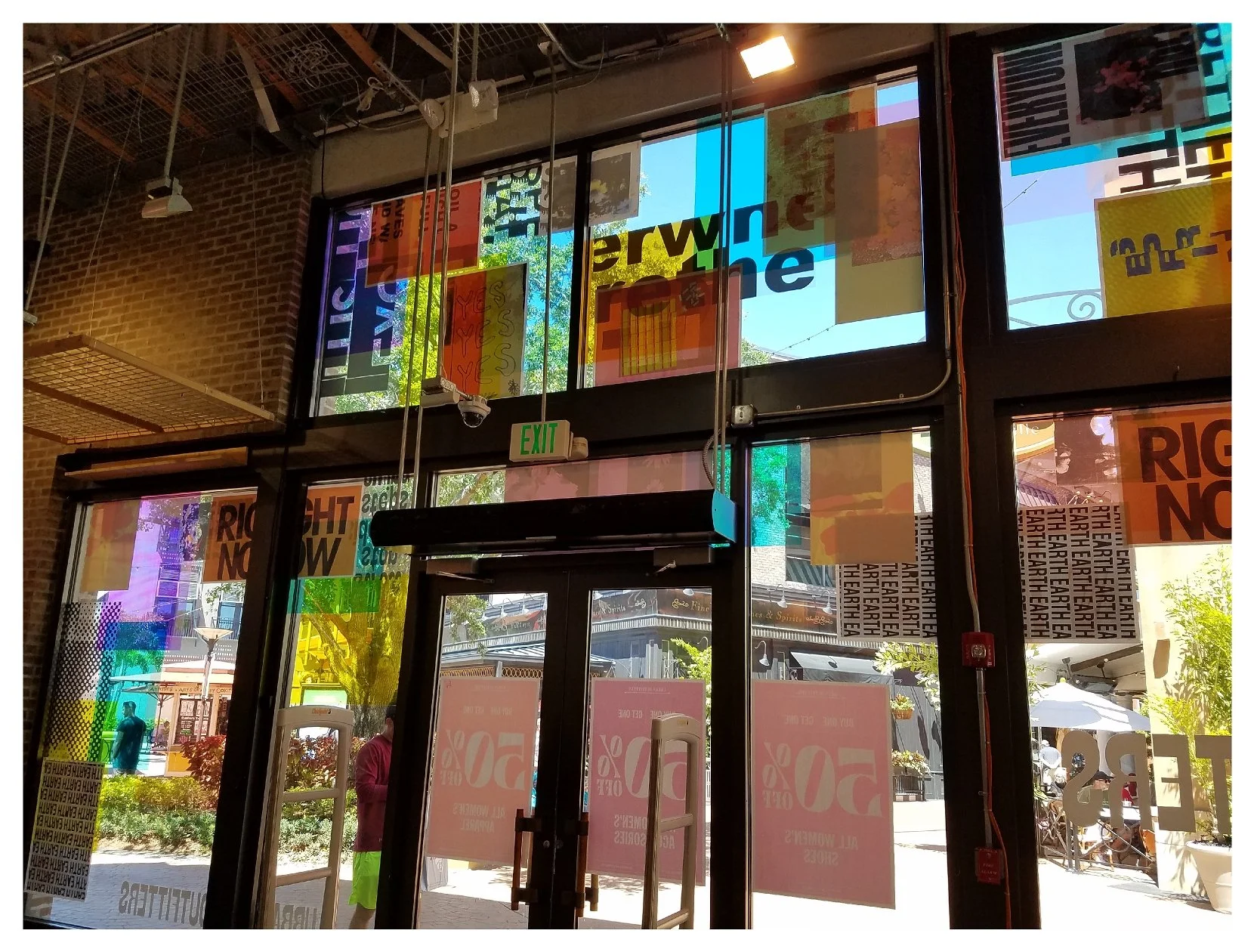 Interior view through glass doors with colorful overlays and text, outdoor scenery visible, featuring sale signs and street scene. colored window film