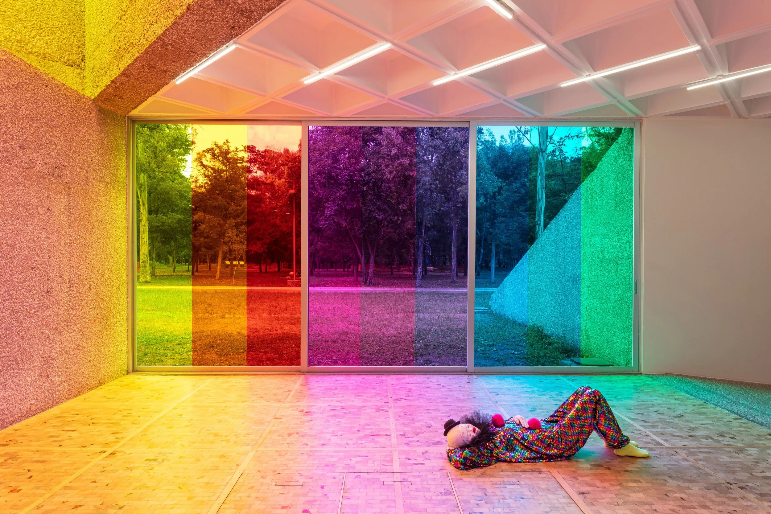 A child lying on the floor wearing a colorful clown costume with a rainbow wig and big red nose, in front of a large window with rainbow-colored glass panes revealing a park with trees outside. The room has a textured yellow and brown wall on the left and a white ceiling with geometric patterns and LED lights.