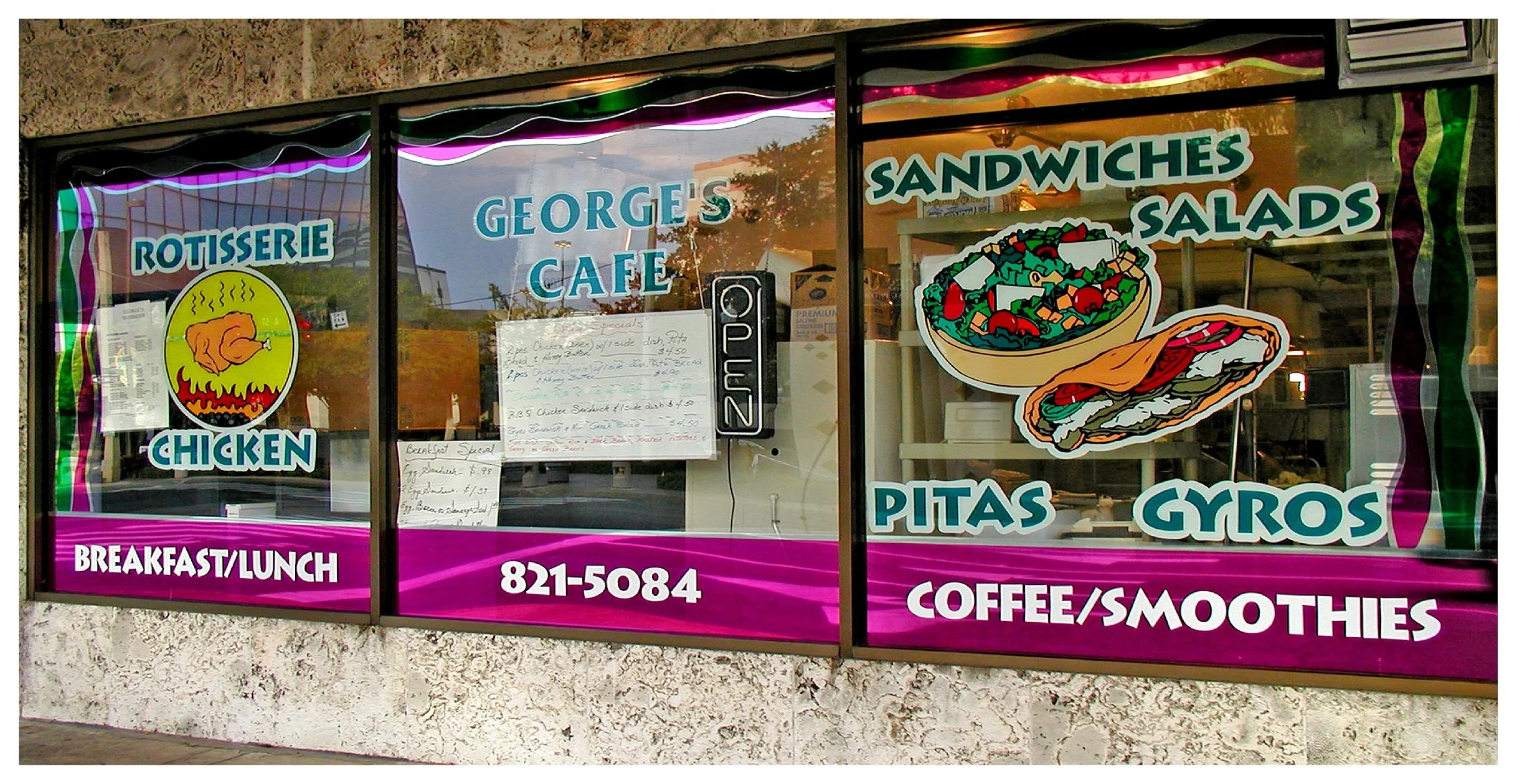 Front window of George's Cafe with signage for rotisserie chicken, sandwiches, salads, breakfast, lunch, pitas, gyros, coffee, and smoothies.