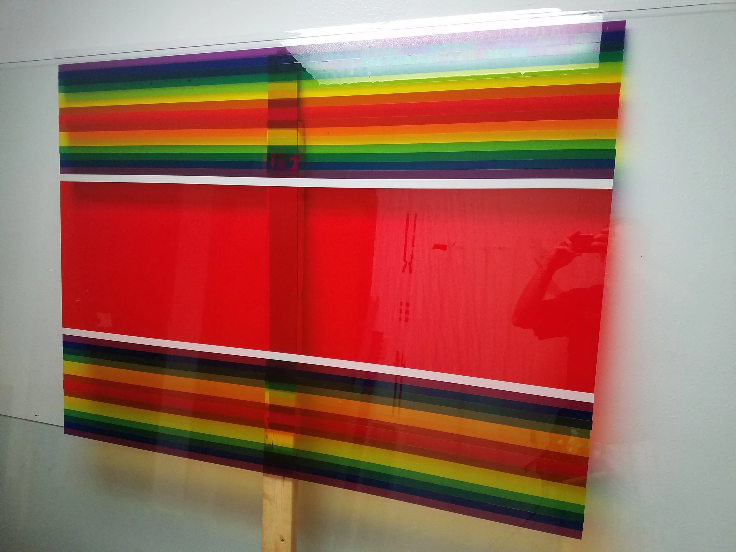 A large, framed artwork with horizontal rainbow-like stripes in purple, blue, green, yellow, orange, and red colors, mounted on a wall. colored window film