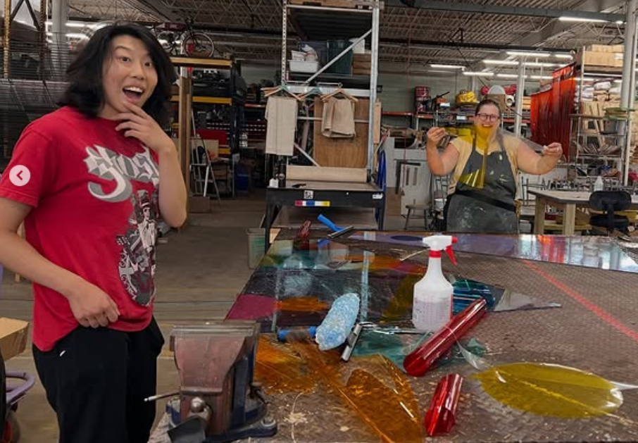 Two women in a workshop with glasswork; one woman in a red shirt with a surprised expression, the other woman behind her holding tools and smiling. colored window film