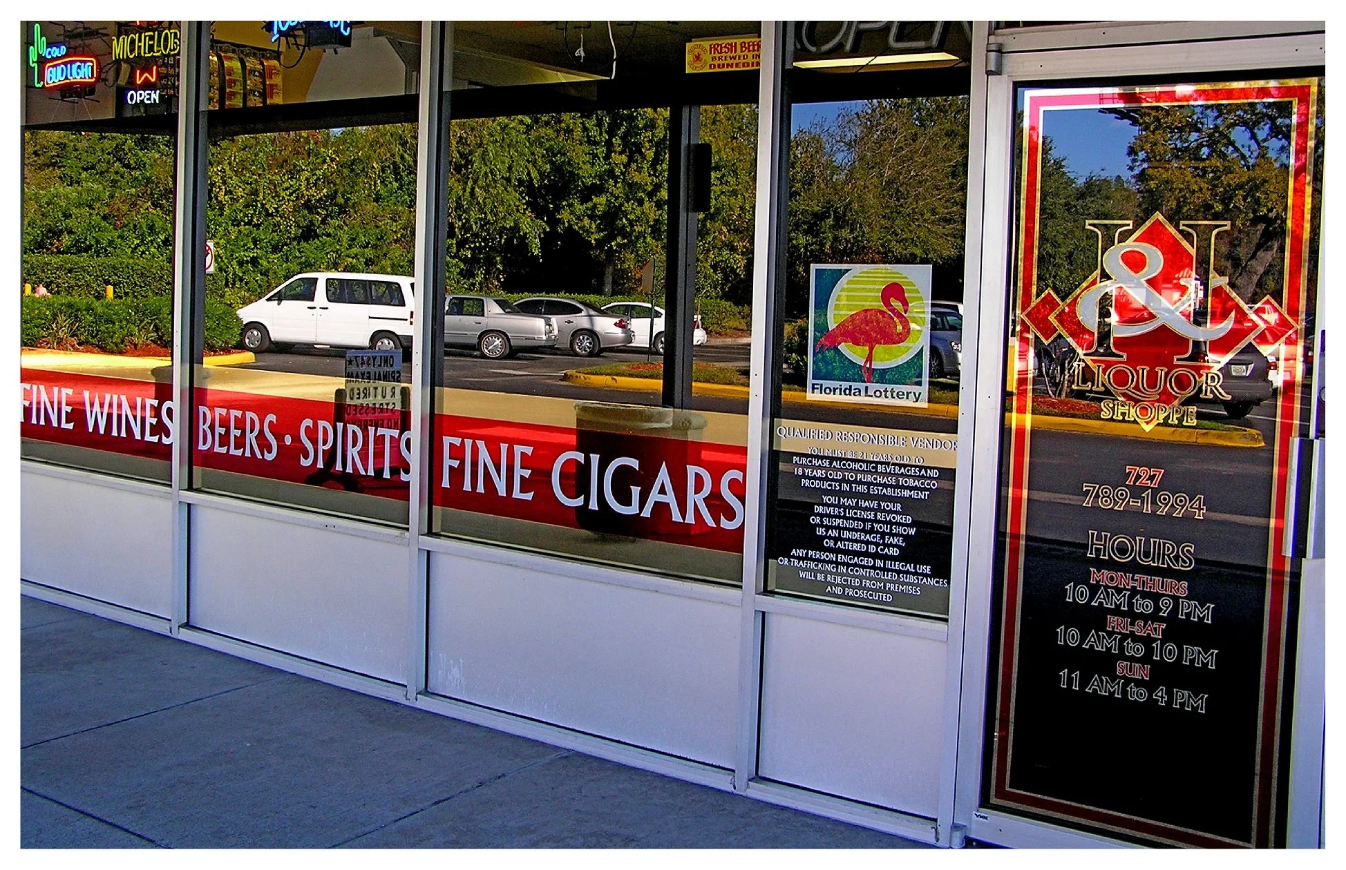 Exterior view of a liquor store with signage for fine wines, beers, spirits, and cigars, featuring Florida Lottery and store hours on the door. colored window film