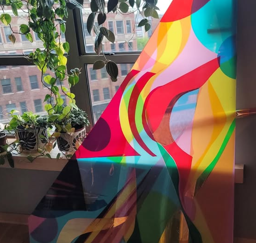 Colorful abstract glass artwork with swirling patterns, placed near a window with potted plants and a cityscape view. colored window film
