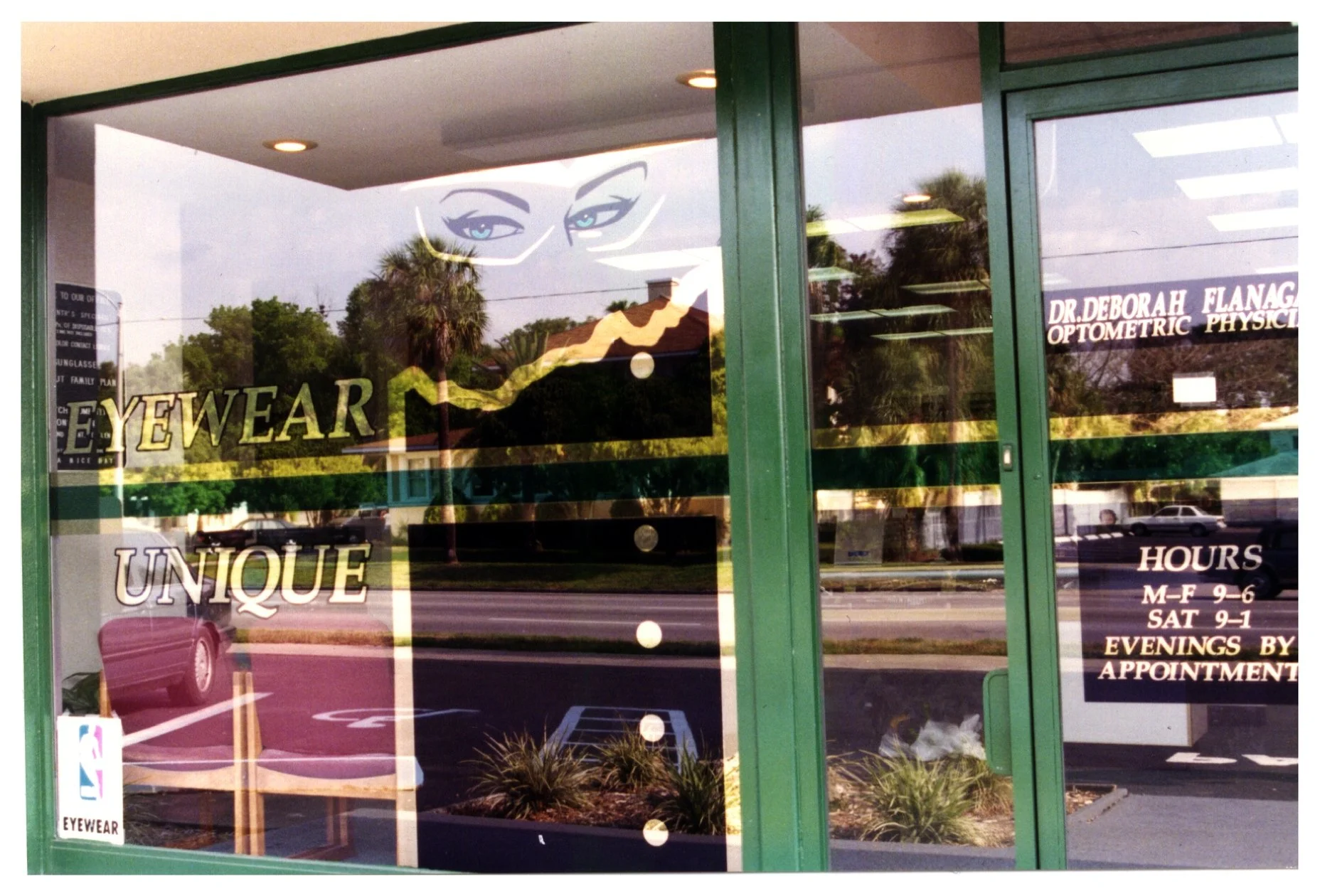 Eyewear store front window with signage, including business hours and optometrist's name, reflecting street and trees. colored window film