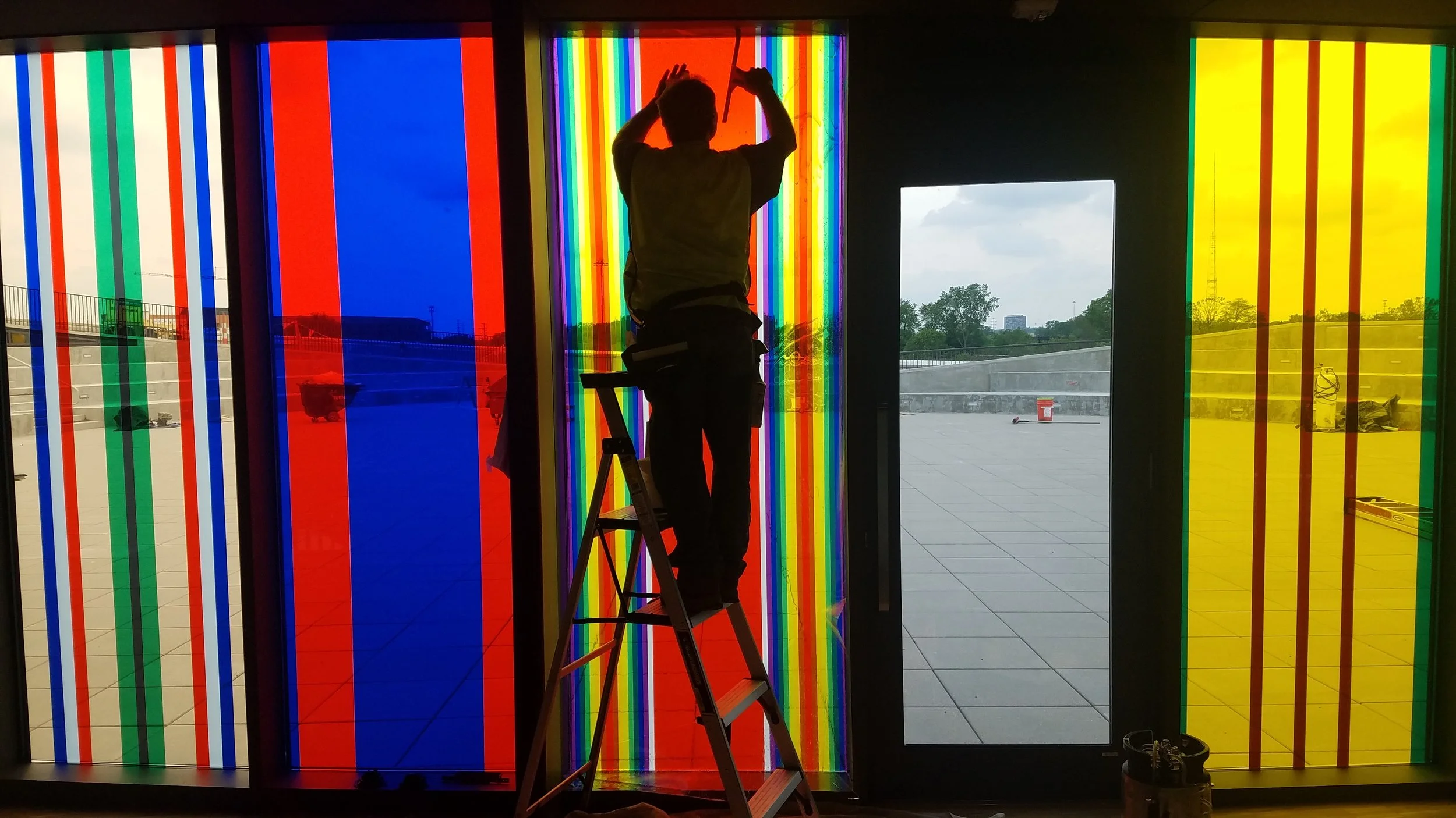Person standing on ladder installing or repairing a colorful, vertically striped glass installation near an open door, with an outdoor view of trees, buildings, and cloudy sky. colored window film