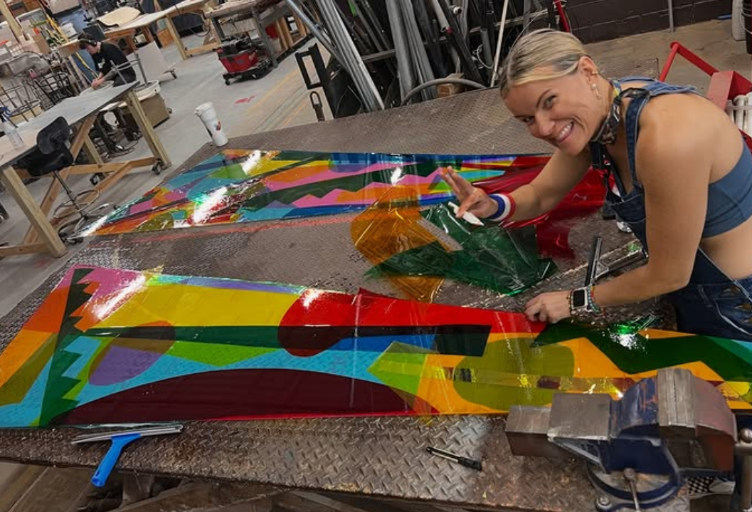 A woman smiling and waving at the camera while working on a colorful, abstract, holographic artwork or sign in a workshop or factory setting. colored window film