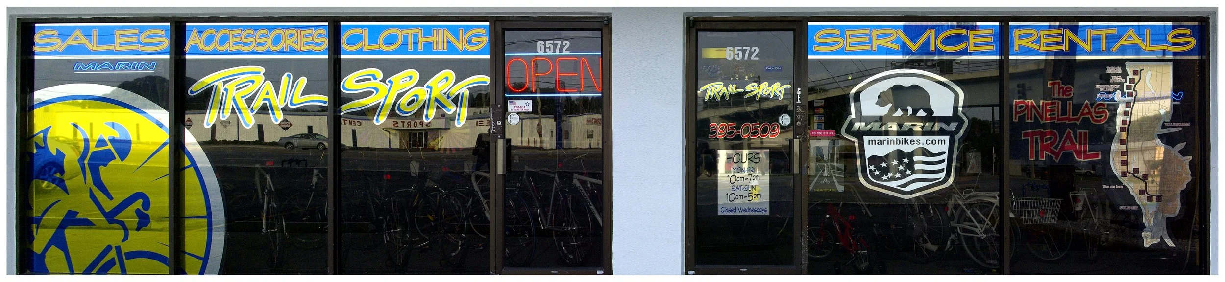 Storefront of Trail Sport bike shop with signs for sales, accessories, clothing, service, and rentals. Features Marin bike logo and open sign. colored window film