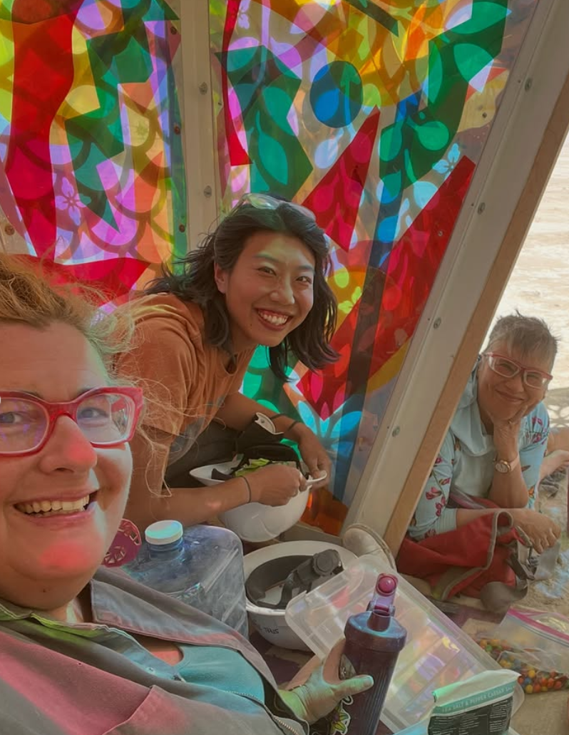 Three smiling women sitting inside a colorful, abstract stained glass window display on a beach. colored window film