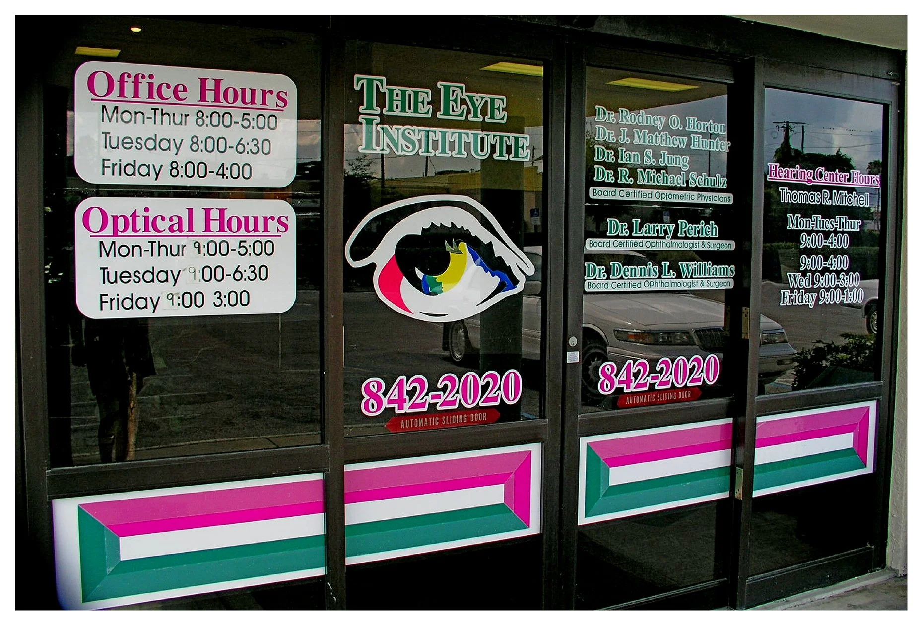 Glass doors of an eye institute with office and optical hours, phone number, doctor names, and a colorful eye graphic. colored window film