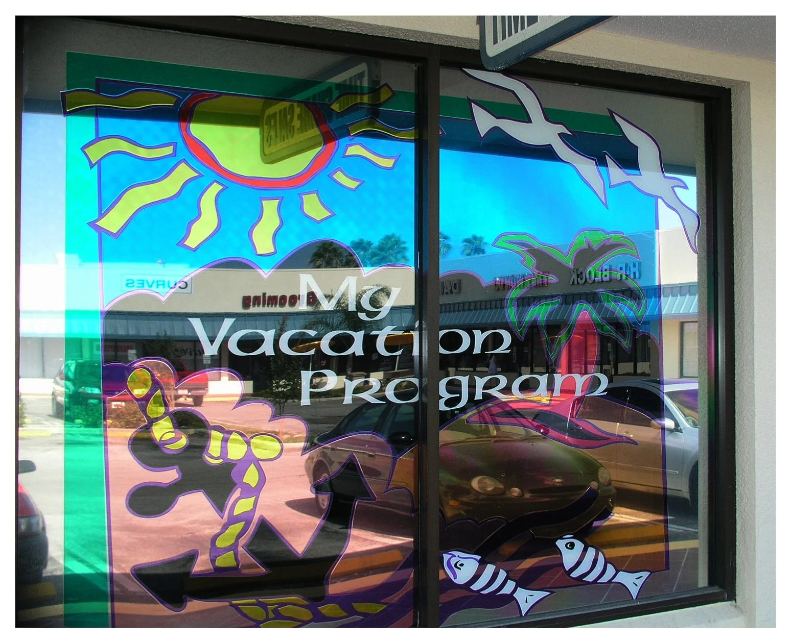 Storefront window with colorful ocean-themed decorations, displaying text 'My Vacation Program', featuring a sun, seagulls, palm trees, anchor, and fish illustrations. colored window film