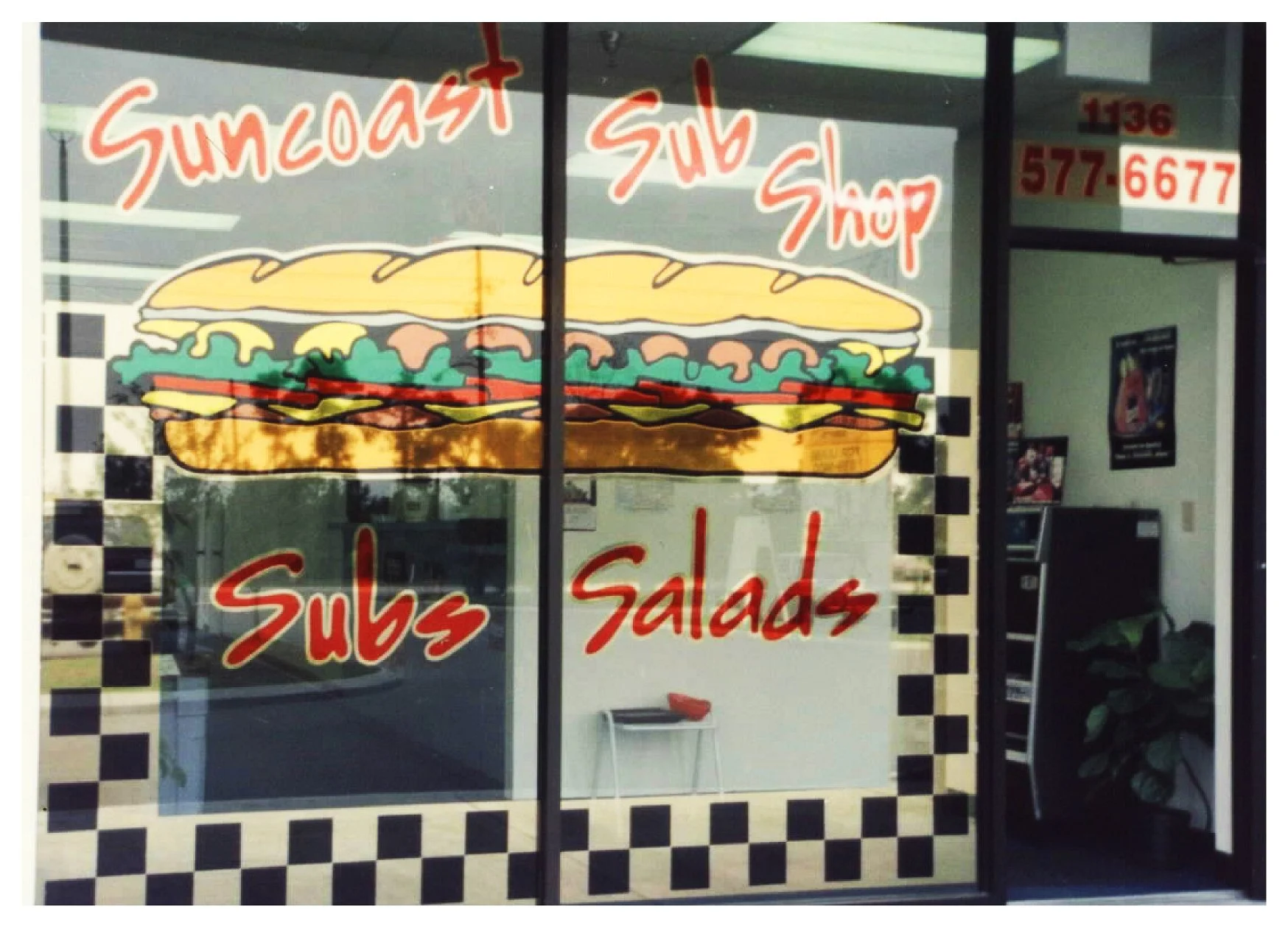 Storefront window of Suncoast Sub Shop featuring a large sandwich graphic, text "Subs Salads," checkered border, and phone number.