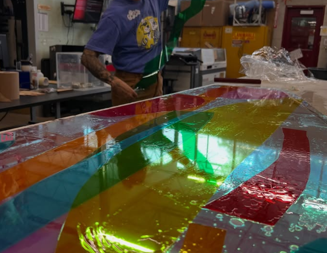 Close-up of a colorful, reflective artwork or display on a table, with a person working in the background in a workshop setting. colored window film