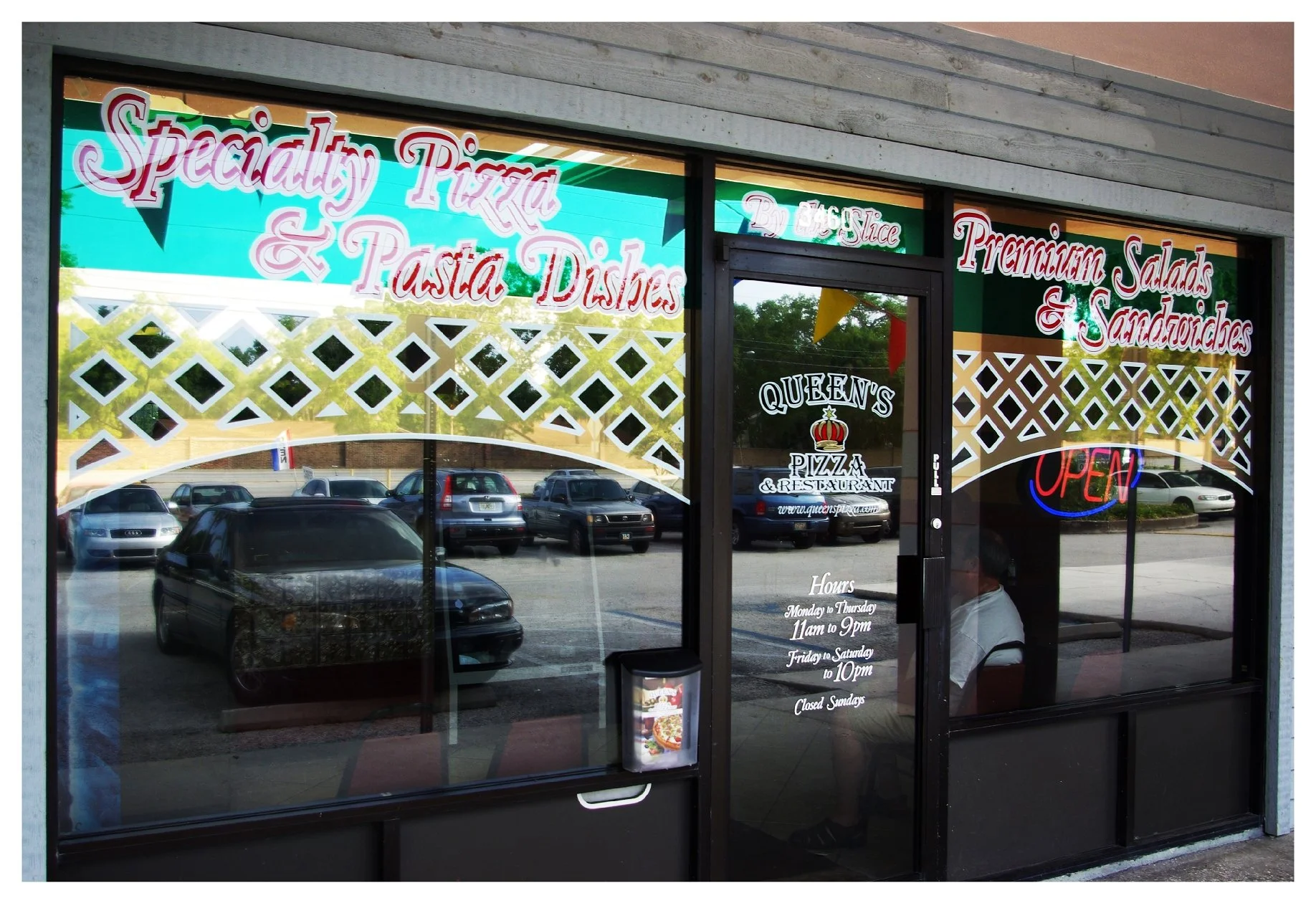 Front window of a pizza and restaurant business with signage for specialty pizza, pasta dishes, premium salads, and sandwiches. The business is open, with listed hours on the door. colored window film