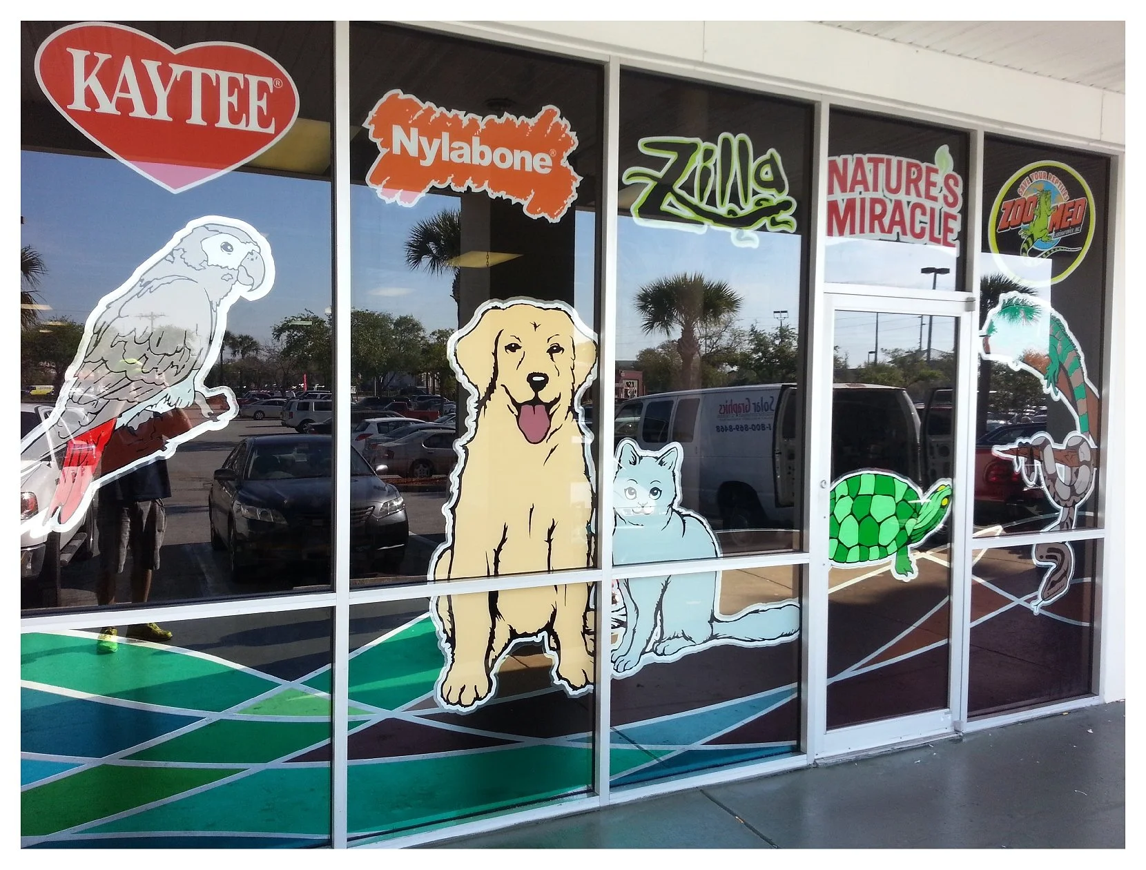 Pet store window with animal decals and brand logos including Kaytee, Nylabone, Zilla, Nature's Miracle, Zoo Med. Animals depicted are a bird, dog, cat, turtle, lizard, and snake. colored window film