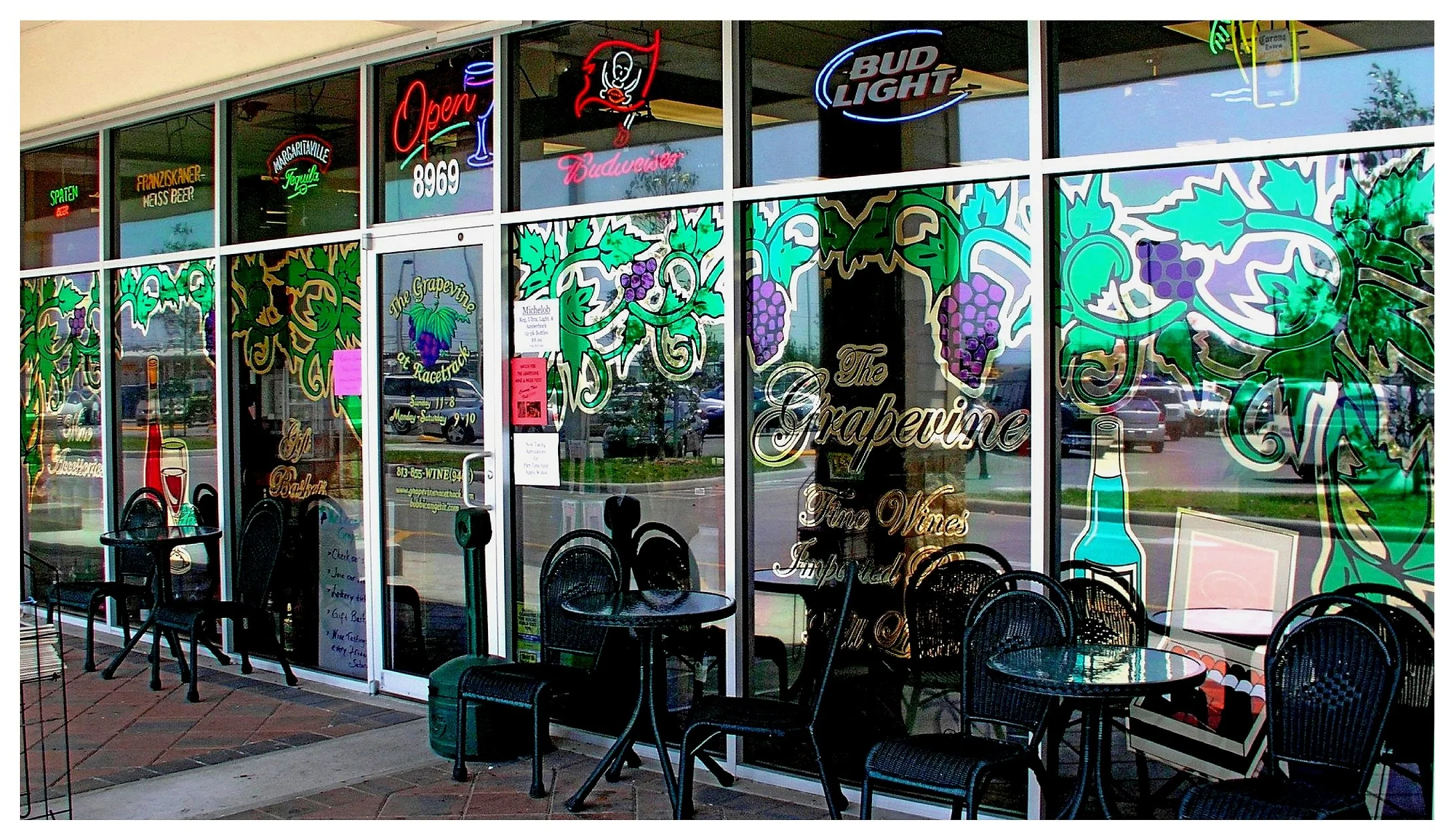 Wine and beer shop storefront with grape-themed decorations, neon signs for various beer brands, and outdoor seating. colored window film