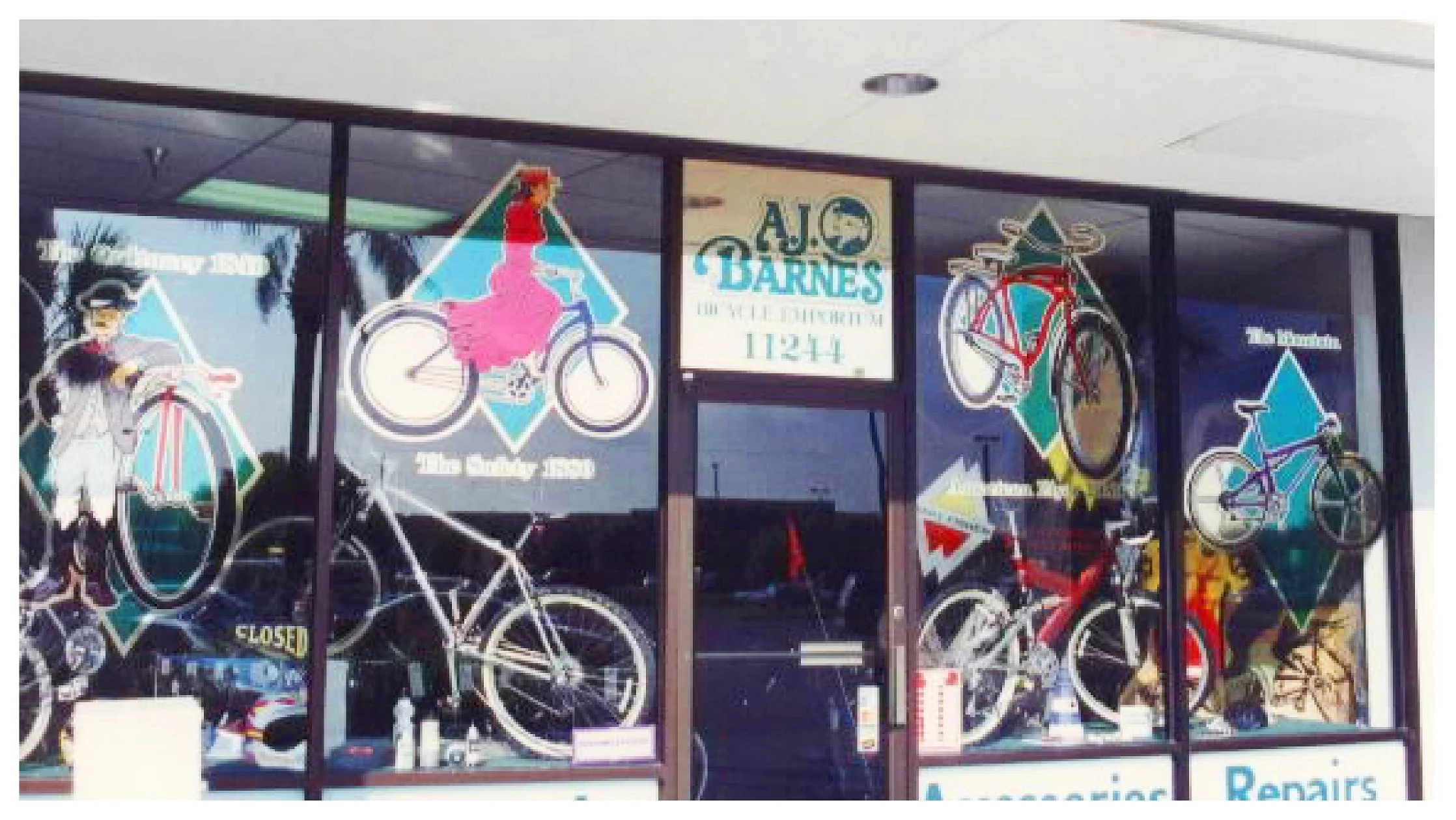 Storefront of A.J. Barnes Bicycle Emporium with window graphics of different bicycles and historical figures riding them. Sign with store name and address '11244' visible. colored window film