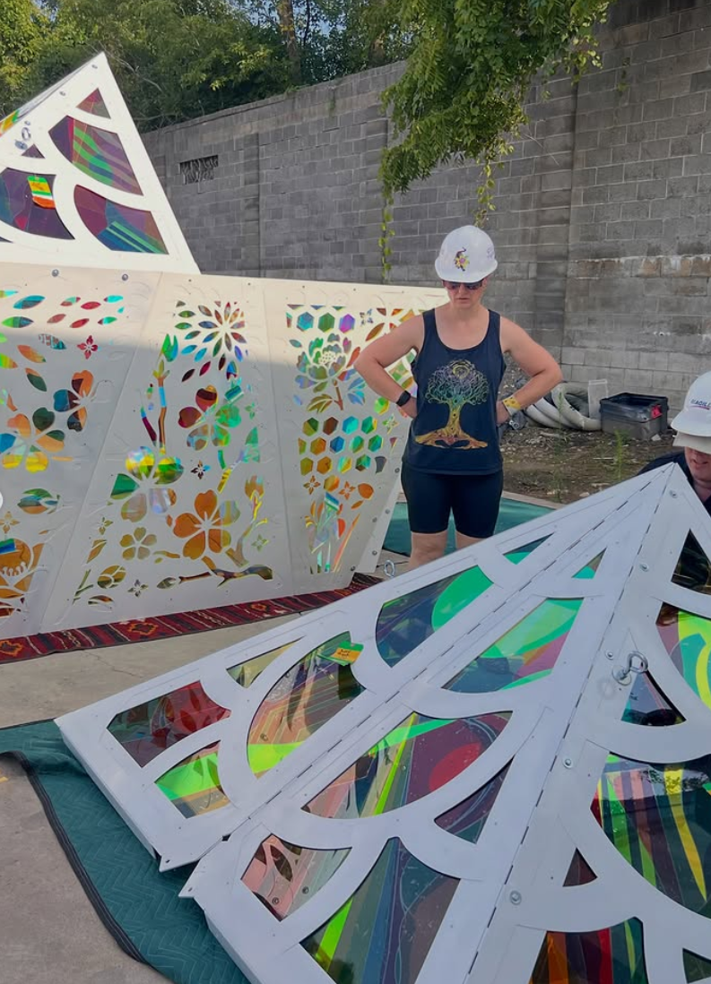 Two women wearing white hard hats and dark clothing are working outdoors near a gray brick wall. They are surrounded by colorful, decorative metal panels with intricate cut-out designs and stained glass insets, laid out on mats. colored window film