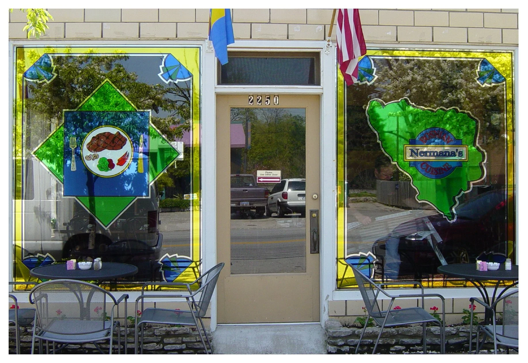 Front of a restaurant with stained glass windows featuring a steak image and "Norma's European Cuisine" logo, outdoor seating with metal chairs and tables, and flags above the door. colored window film