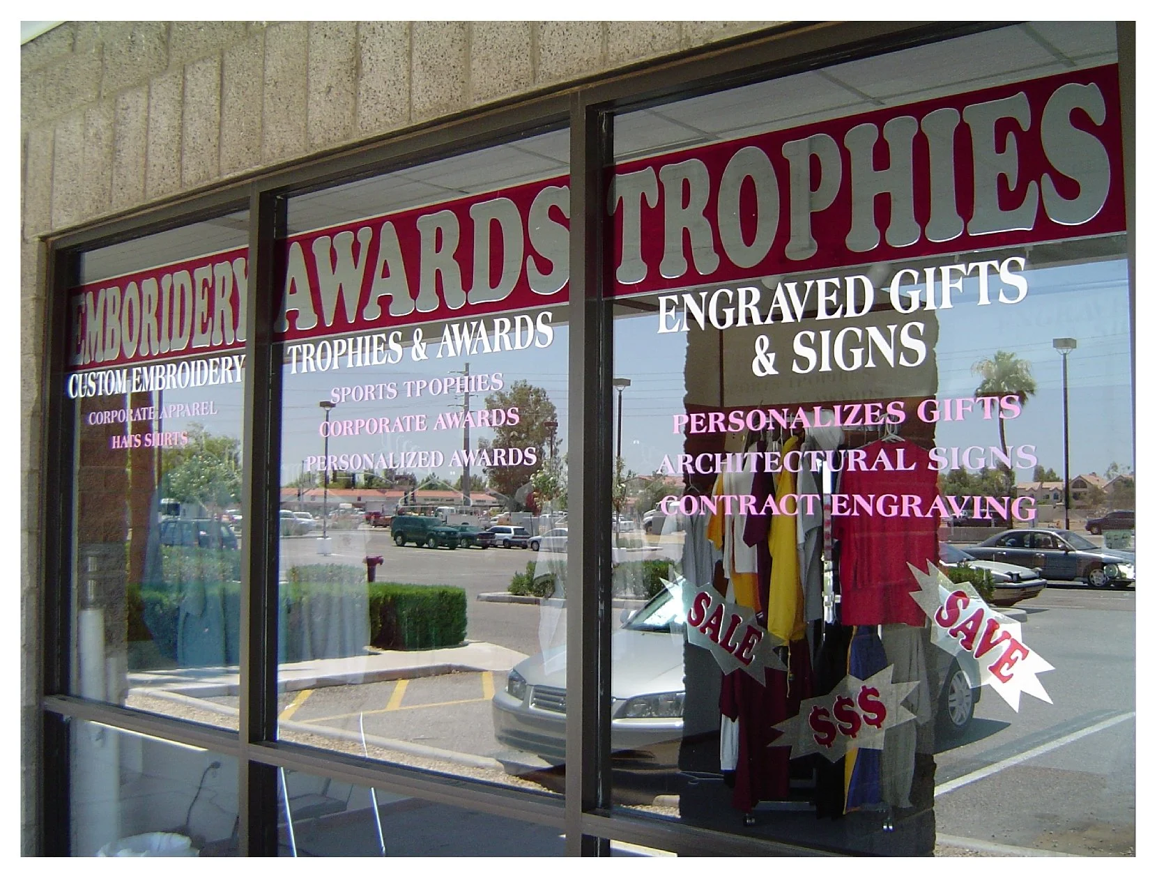 Storefront window displaying text advertising embroidery, awards, trophies, engraved gifts and signs, with reflections of parked cars and trees. colored window film