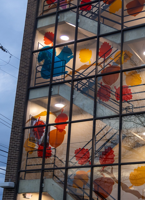 A multi-story building with a glass exterior featuring colorful leaf and branch decals inside, viewed from outside during the evening. colored window film