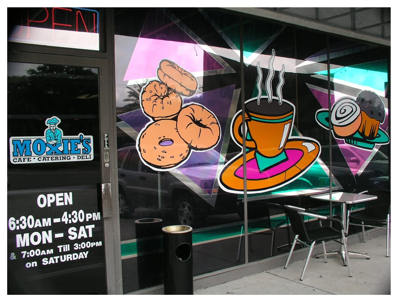 Exterior of cafe with colorful graphic of coffee, bagels, and pastry on window; signage indicates hours and name "MOXIE'S Cafe, Catering, Deli"; outdoor seating with table and chairs.