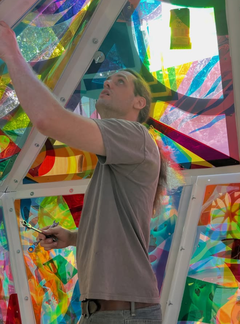 A man in a gray t-shirt climbing inside an interactive, multicolored geometric art installation, looking upward while holding a small tool. colored window film