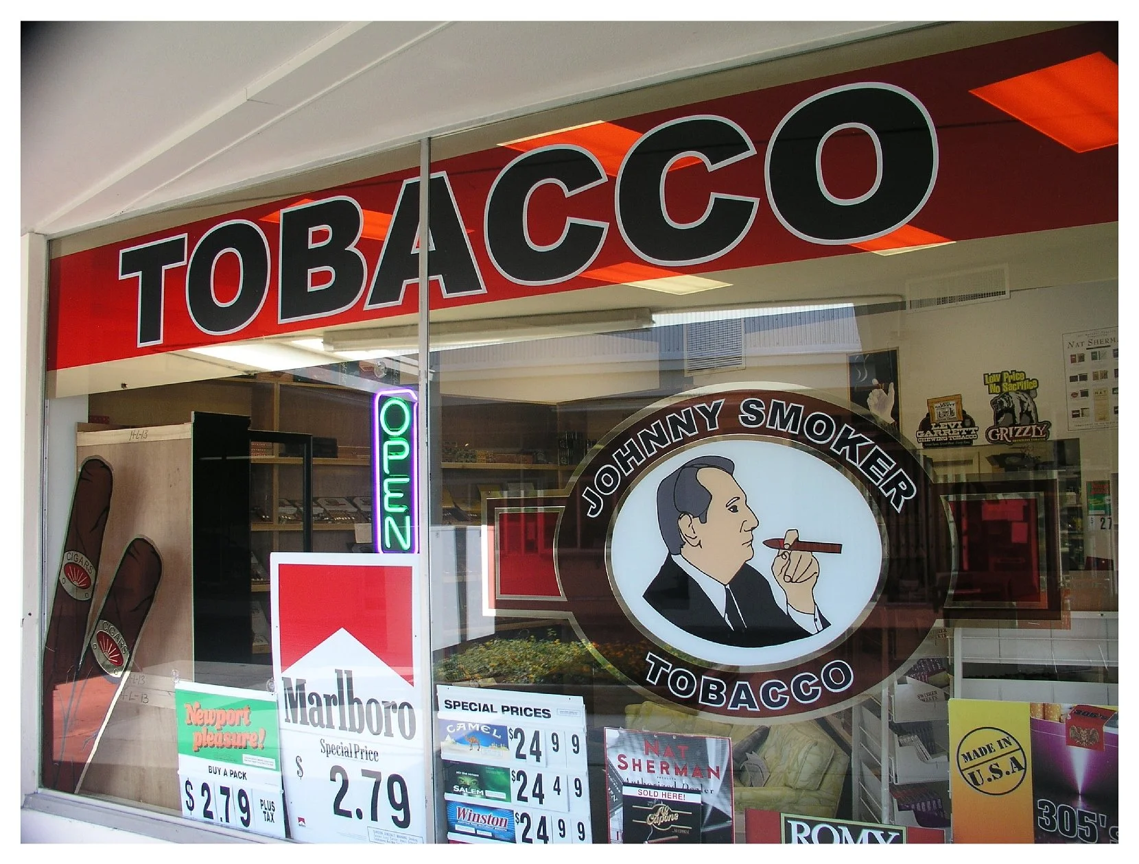 Storefront of a tobacco shop with "Tobacco" sign, "Johnny Smoker" logo, neon "Open" sign, and various cigarette brand advertisements including Marlboro. colored window film