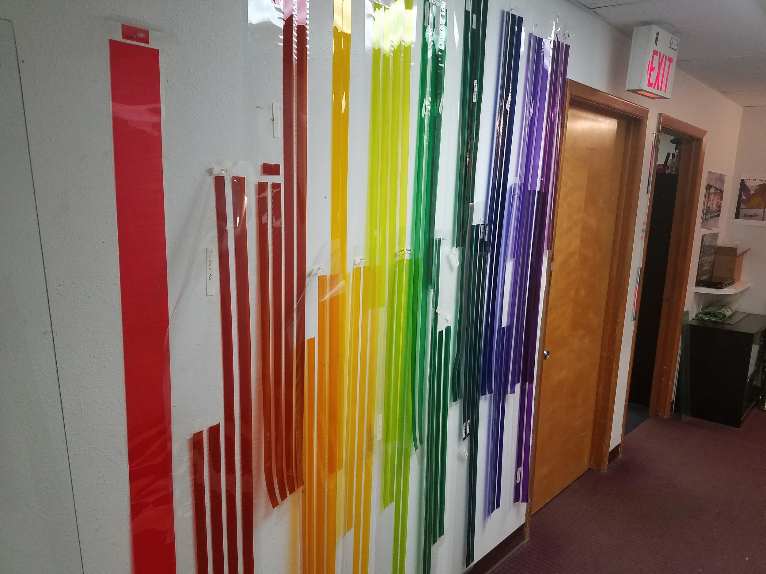 A wall display of long, narrow, colored film strips arranged in a rainbow order from red to purple. colored window film