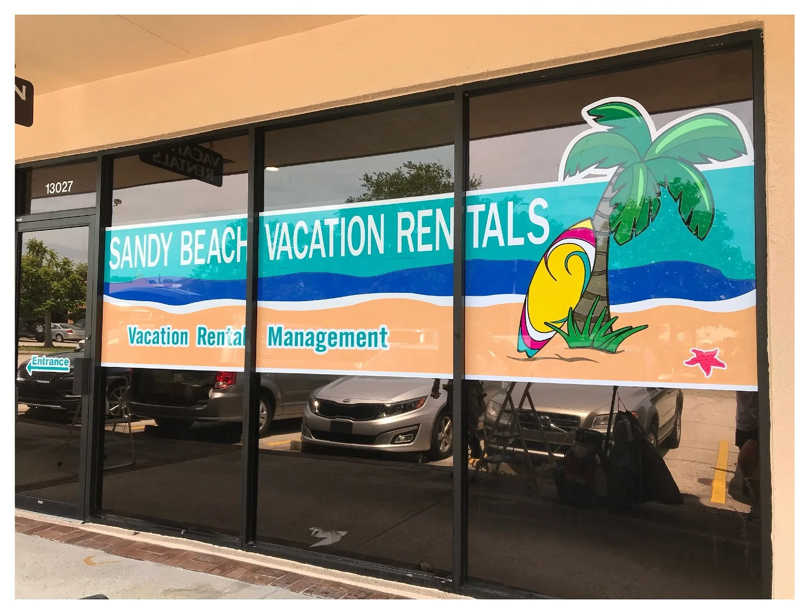Window display of Sandy Beach Vacation Rentals with graphics of a palm tree and surfboard. colored window film