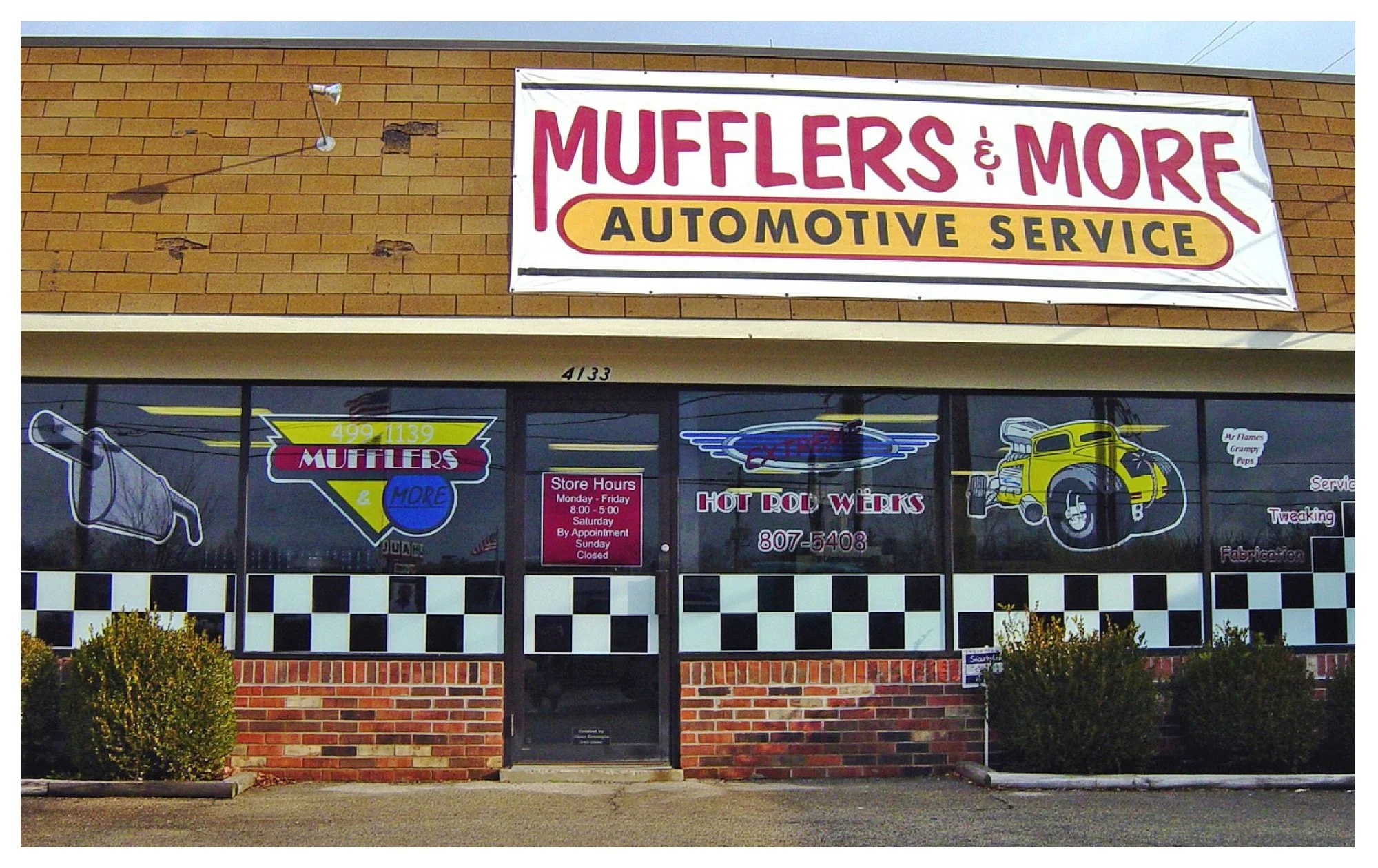 Front of automotive service shop with signs for 'Mufflers & More' and 'Hot Rod Werks' on the windows.