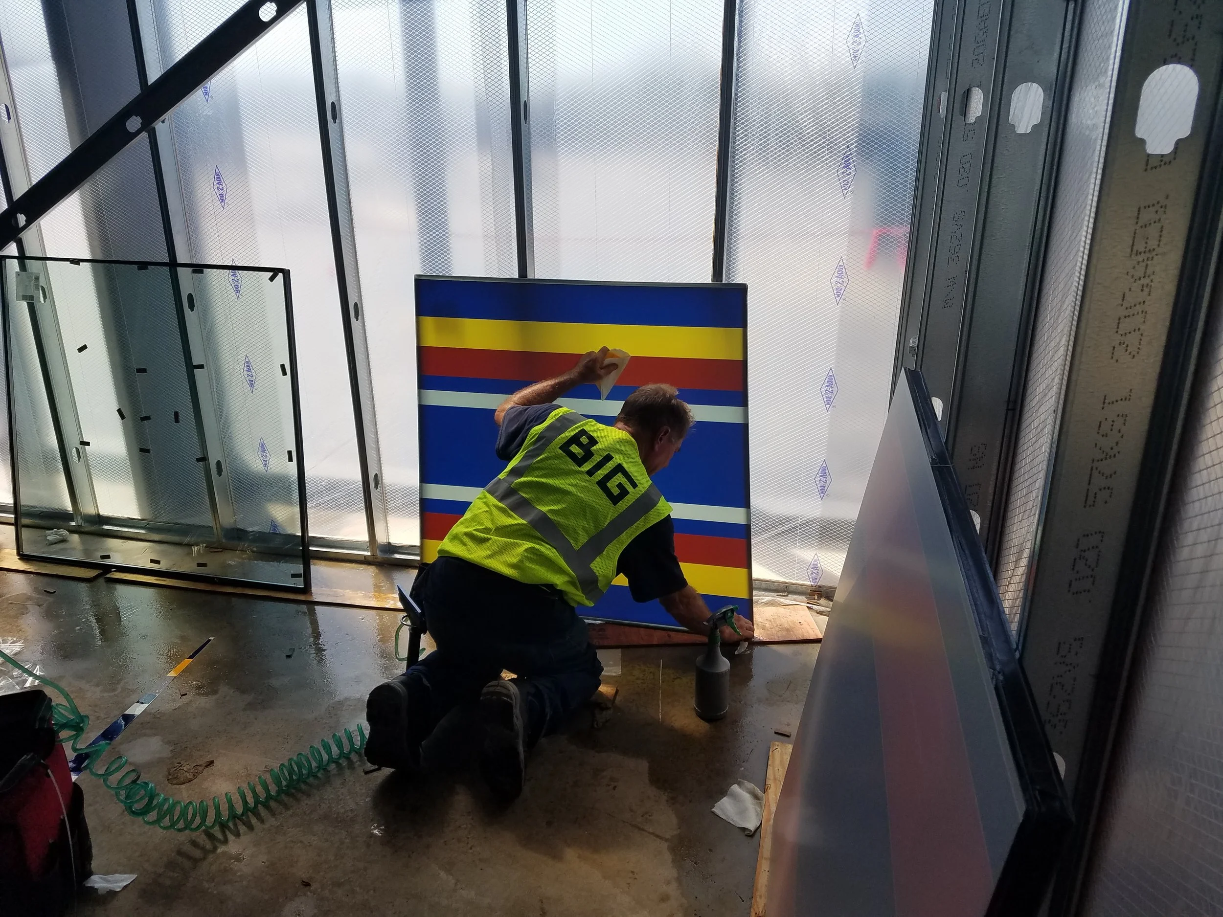 Worker installing a large sign with horizontal stripes in yellow, red, and blue at a construction site inside a building under renovation. Installing color window tint graphics.