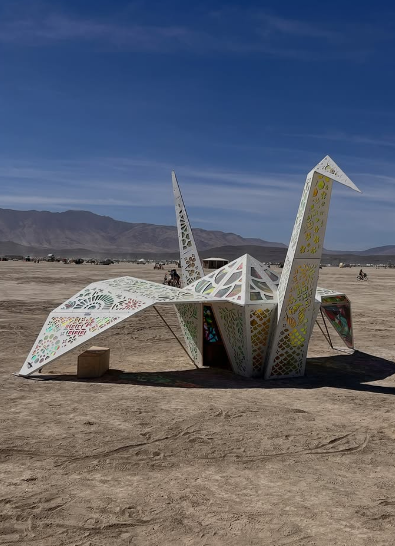 Art installation resembling a futuristic sculpture covered in colored window film in a desert landscape with mountains and people riding bikes in the background.