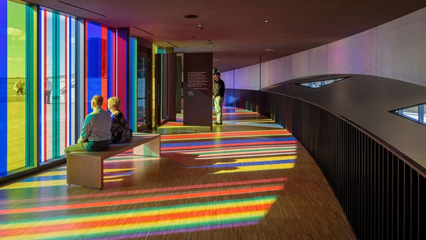 Two people sitting on a bench inside a modern building, overlooking a colorful, rainbow-patterned light display on the floor and wall made by sunlight passing through vibrantly colored window film panels.