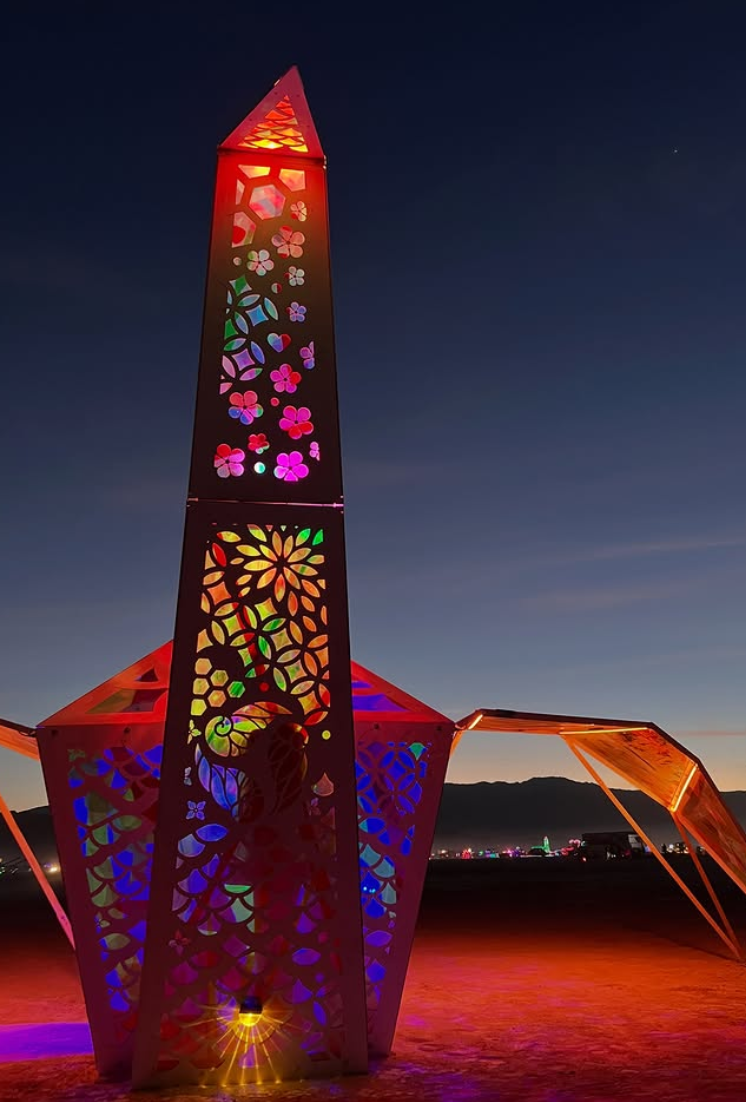 Colorful illuminated sculpture covered with colored window film, set against a twilight sky in a desert landscape.
